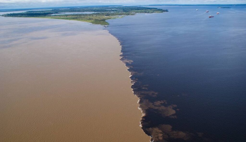 An aerial photograph showing the exact meeting point of two massive rivers, where dark blue water meets turbid, light brown water, creating a sharply defined, swirling line.