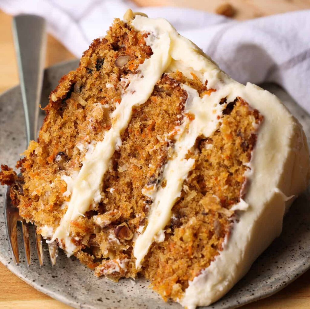A thick slice of three-layer carrot cake with cream cheese frosting and visible walnuts on a speckled grey plate.Soft Decadent Carrot Cake