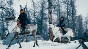 Two children riding on the backs of reindeer through a snow-covered forest with a traditional conical tent in the background.