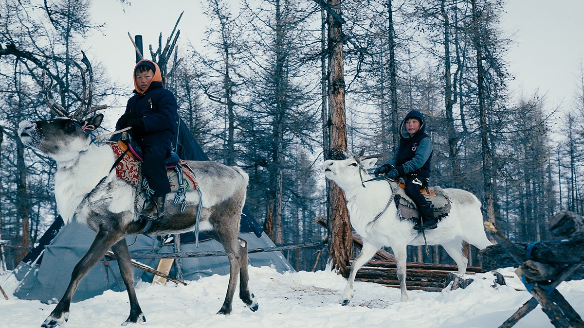 Two children riding on the backs of reindeer through a snow-covered forest with a traditional conical tent in the background.