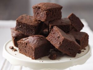 A tall stack of thick, square-cut chocolate brownies with a visible fudgy crumb and light peanut butter swirls, displayed on a white ceramic cake stand.