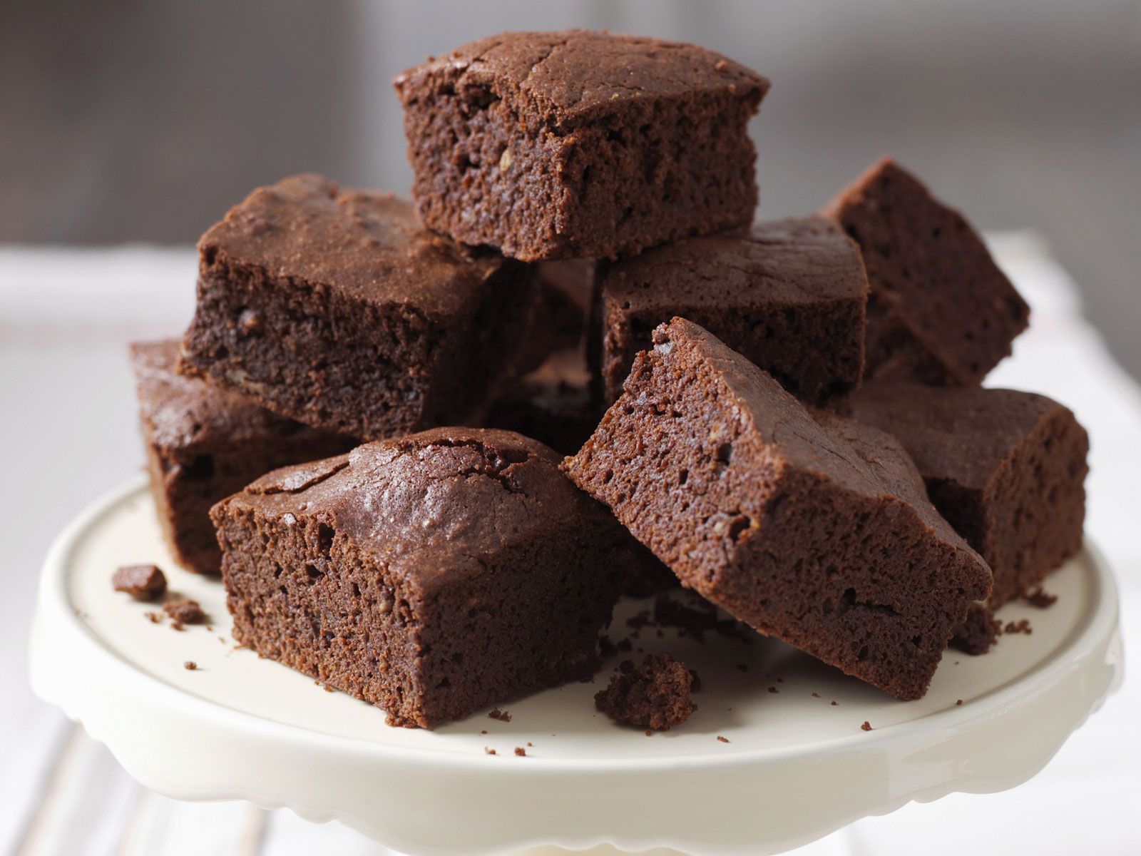 A tall stack of thick, square-cut chocolate brownies with a visible fudgy crumb and light peanut butter swirls, displayed on a white ceramic cake stand.