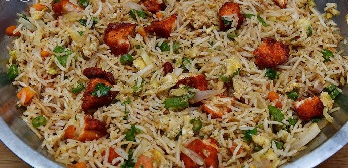 A close-up shot of Indian-style chicken fried rice in a metal pan, featuring long-grain basmati rice, fried chicken pieces, scrambled eggs, and green onions.