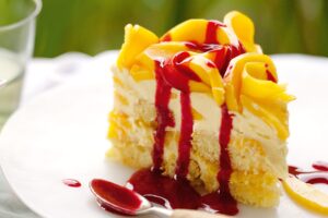 A slice of layered mango tiramisu cake on a white plate, topped with fresh mango slices and drizzled with a bright red raspberry coulis.