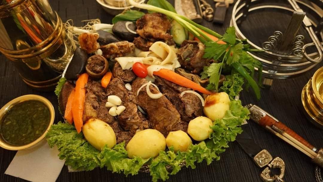 A large traditional Mongolian platter filled with slow-cooked mutton, steamed potatoes, and carrots, garnished with green onions and lettuce, surrounded by traditional metalware and a side of dipping sauce.
