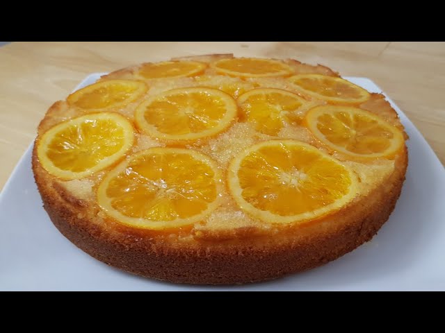 A round, golden-brown orange upside-down cake topped with thin, glazed orange slices arranged in a circular pattern on a white plate.