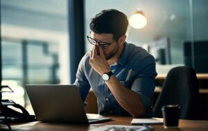 A man in a blue shirt and glasses sitting at a desk with a laptop, pinching the bridge of his nose in a gesture of stress or exhaustion.