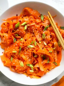 A white bowl filled with wide, ribboned carrot shavings topped with sliced green scallions, white sesame seeds, and red chili flakes, shown with wooden chopsticks.