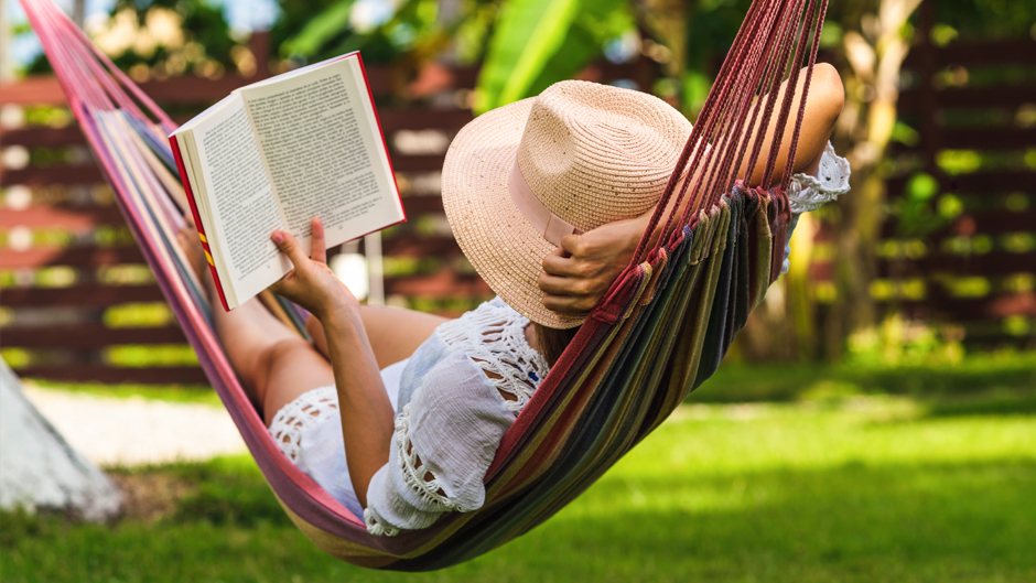 A person wearing a sun hat and white crochet dress relaxes in a colorful striped hammock while reading a book in a lush, green outdoor setting.